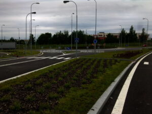 Zakřivená silnice s několika pouličními lampami, přechody a dopravními značkami v příměstské oblasti; uprostřed silnice je vysázena tráva a malé keře a v pozadí jsou pod zataženou oblohou vidět budovy a stromy.