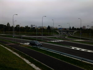 U prázdné víceproudé silnice s několika pouličními lampami a dopravním značením pod zataženou oblohou stojí tmavé auto. V pozadí jsou stromy, tráva a vzdálené budovy.