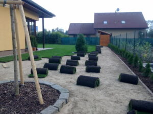 Na písečné cestě vedle domu jsou položeny role drnů připravené k rozbalení. Malý stromek je podepřen dřevěnými kůly v mulčované oblasti. V pozadí stojí další dům a kůlna.