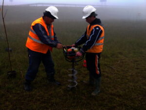 Dva pracovníci v oranžových bezpečnostních vestách a bílých přilbách vrtají mechanickým vrtákem do země na travnatém, mlhavém venkovním prostranství.
