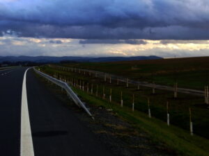 Křivolaká silnice vede podél travnatého pole s mladými stromky a zábradlím pod dramatickou zamračenou oblohou, na obzoru jsou vidět vzdálené kopce.