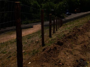 Drátěný plot s dřevěnými sloupky vede podél prašného svahu vedle štěrkové cesty. V pozadí jsou vidět stromy a stavební vozidla.