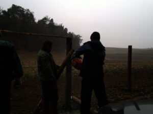 Dva lidé jsou venku a pomocí motorové pily řežou dřevěný sloup ve venkovské krajině se stromy a poli v pozadí pod zataženou oblohou.