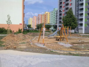 Malé dětské hřiště se skluzavkou a prolézačkami je ve výstavbě, obklopené hromadami hlíny a výstražnou páskou, s barevnými bytovými komplexy v pozadí.