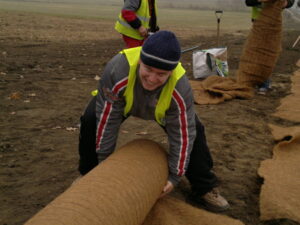 Osoba ve žluté bezpečnostní vestě a modré pletené čepici se usmívá při rozbalování velké role kokosové rohože na holé zemi, v pozadí pracují další lidé.