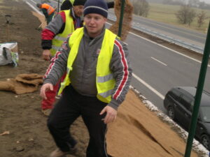 Muž ve žluté bezpečnostní vestě a zimní čepici pracuje s dalším pracovníkem na náspu u silnice. Na půdu vedle dálnice rozprostírají hnědou podložku. V pozadí je vidět auto a lopaty.