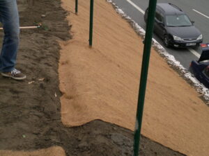 Osoba stojí na půdě vedle svahu pokrytého protierozní rohoží vedle silnice s projíždějícími auty. Podél svahu jsou rozmístěny zelené sloupky.