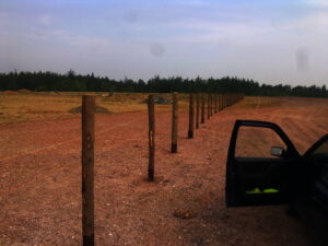 Na pustém kamenitém poli s lesem v pozadí stojí řada dřevěných plotových sloupků. V pravé části snímku jsou vidět otevřené dveře černého vozidla.