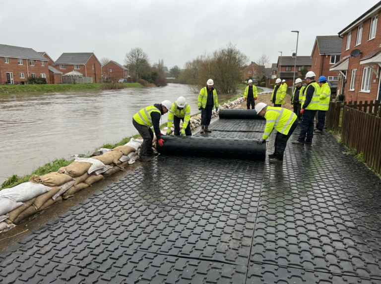 Dělníci ve vestách s vysokou viditelností a přilbách instalují u řeky protipovodňové zábrany, jejichž okraj lemují pytle s pískem. Nedaleko jsou domy a půda je mokrá po nedávném dešti.