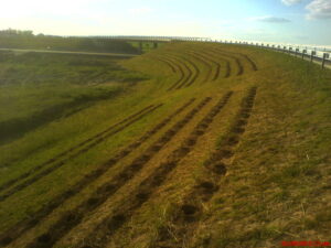 Na travnatém svahu vedle silnice s mostem v pozadí jsou pod jasnou modrou oblohou vykopány řady kruhových jam.