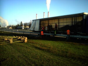 Dělníci v oranžových a žlutých bezpečnostních vestách stojí u otevřeného nákladního auta u silnice, v pozadí jsou vidět kouřící komíny a v popředí leží na trávě svázané mladé stromky.