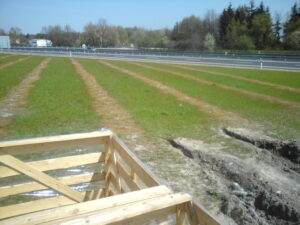 Travnaté pole s rovnoměrně rozmístěnými řádky, ohraničené silnicí a stromy v pozadí. V popředí stojí dřevěná konstrukce poblíž plácku holé půdy a mělkého příkopu, což naznačuje nedávné chemické ošetření trávníku.