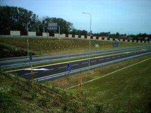 Nově postavená dálnice s více jízdními pruhy, zřetelným dopravním značením a modrými dopravními značkami. Po stranách jsou travou porostlé náspy a mladé stromy, v pozadí jsou vidět protihlukové stěny a domy.