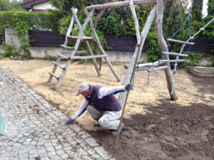 Osoba v pracovním oděvu a rukavicích klečí na zemi vedle dětského hřiště a pracuje na okraji dlážděné cesty vedle dřevěné prolézačky. Plocha je obklopena zelení a dřevěným plotem.