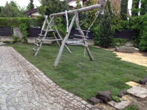 Dvůr s nově položeným trávníkem, dřevěnou houpačkou s žebříkem a houpačkou z pneumatik, pískovištěm a dlážděnou cestou lemující travnatou plochu.