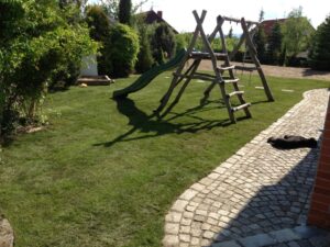 Na dvorku se zelenou trávou je dřevěná houpačka a skluzavka. Vedle ní vede zakřivená kamenná cesta a na trávě u ní leží černý pes. Dvůr lemují stromy a keře.