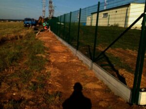 Podél polní cesty vede kovový plot a v jeho blízkosti jsou vidět stavební dělníci a technika včetně modrého nákladního auta. V pozadí pod jasnou oblohou stojí bílá budova a věže inženýrských sítí.