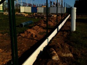 Staveniště s částečně instalovaným kovovým plotem a betonovou podezdívkou, roztroušenými hromadami hlíny a stavebním materiálem na zemi. V pozadí jsou vidět průmyslové stavby a oplocení.