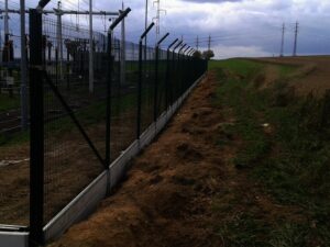 Vysoký kovový plot s ostnatým drátem na vrcholu vede podél elektrické rozvodny a polní cesty, v pozadí je elektrické vedení a zatažená obloha.