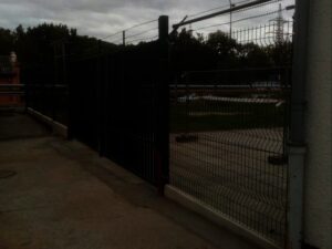 Temný, stínový obraz kovového plotu a brány vedle betonové cesty se stromy a zataženou oblohou v pozadí. Osvětlení je tlumené, takže je obtížné jasně rozeznat detaily.