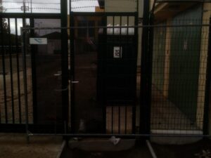 Přístup k přenosné toaletě za nimi blokuje zamčená brána a drátěný plot. Scéna se zdá být tmavá a nachází se venku, poblíž budovy a zpevněné plochy.