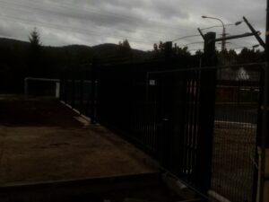 Tmavá, zamračená venkovní scéna zobrazující vysoký kovový plot s ostnatým drátem nahoře, zavřenou bránu a dlážděnou zem. V pozadí jsou pod zataženou oblohou vidět stromy a kopce.