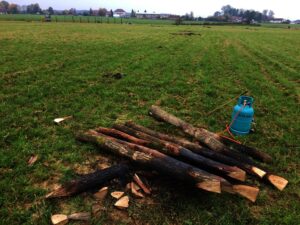Na travnatém poli vedle modré nádrže na propan leží několik dřevěných klád se špičatými konci. V pozadí jsou stromy, domy a vzdálené budovy pod zataženou oblohou.