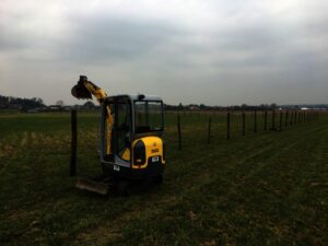 Na travnatém poli u řady nově instalovaných plotových sloupků parkuje pod zamračenou oblohou malý žlutý bagr. V dálce se na obzoru rýsují domy a stromy.