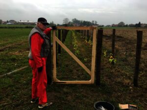 Osoba v červeném oděvu a šedé vestě stojí u dřevěné branky připevněné k plotu na poli, podél kterého rostou malé rostliny pod zataženou oblohou.