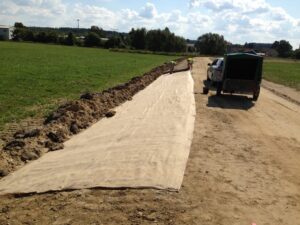 Dva dělníci pokládají velkou plachtu látky nebo geotextilie podél polní cesty na venkově vedle zelené dodávky, v pozadí jsou vidět pole a stromy pod částečně zataženou oblohou.