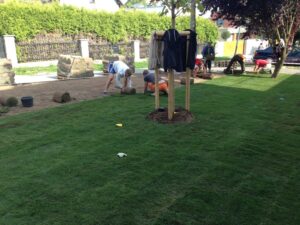 Několik lidí pokládá na dvoře role drnů a společně pracují na instalaci nového trávníku. V popředí stojí mladý stromek s pověšenou bundou a na zemi leží rozházené zahradnické nářadí.