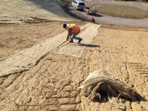 Pracovník v reflexní vestě rozbaluje velkou rohož na holém svahu, pravděpodobně kvůli ochraně proti erozi. Nedaleko leží svázaná rohož a v pozadí na štěrkové cestě jsou dva lidé a vozidlo.