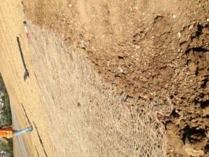 Pracovník v ochranné vestě stojí na svažitém kopci, kde byla na holou půdu částečně položena protierozní rohož. Nedaleko jsou roztroušeny nástroje.