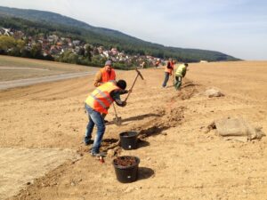 Dělníci v oranžových bezpečnostních vestách sázejí vegetaci na neúrodné svažité ploše poblíž vesnice pomocí lopat a košů se sazenicemi pod částečně zataženou oblohou s kopci a domy v pozadí.