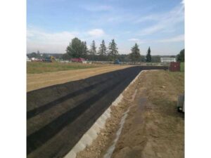 Do dálky se táhne nově vydlážděná silnice ve výstavbě, lemovaná hlínou a trávou, v pozadí jsou vidět stavební vozidla a stromy pod částečně zataženou oblohou.