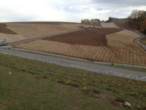 Svažitá krajina s úseky čerstvě obdělané půdy a nově vysazenou vegetací, kterou protínají štěrkové cesty a odvodňovací kanál vyložený kameny; v pozadí je vidět několik vozidel a budov.