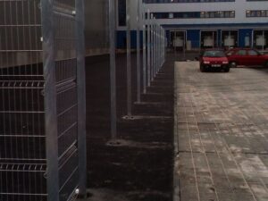 Řada kovových plotových sloupků vede podél zpevněné plochy se zaparkovanými auty v blízkosti velké průmyslové budovy s několika nakládacími rampami a modrými vraty v pozadí.