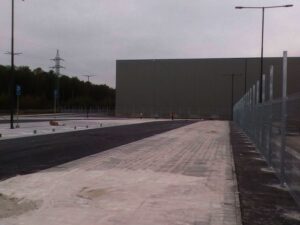 Prázdné, čerstvě vydlážděné parkoviště s pouličním osvětlením, vysoký drátěný plot vpravo a velká hladká budova v pozadí pod šedou oblohou. Nejsou zde přítomna žádná vozidla.