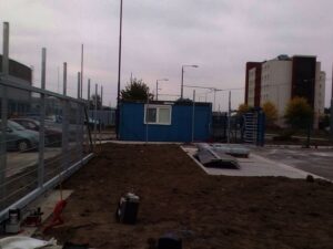 Staveniště s kusem holé půdy, kovovým oplocením, roztroušeným nářadím a modrou přenosnou budovou v pozadí. Pod zataženou oblohou je vidět několik aut a několikapatrová budova.