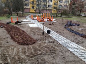 Nově vybudovaná parková plocha s dlážděným chodníkem, oranžovým zábradlím, malým mostem, čerstvě vysazenými záhony, zahradnickým nářadím a nedalekými bytovými komplexy v pozadí.