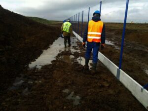 Dva stavební dělníci v ochranných pomůckách kráčejí po blátivé, podmáčené cestě vedle modrého kovového plotu pod zamračenou oblohou. Půda je nerovná a mokrá a oba pracovníci mají na sobě holínky a viditelné vesty.