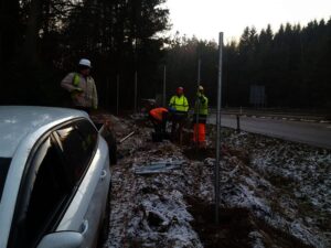 Čtyři stavební dělníci v ochranných pomůckách pracují u silnice s kovovými tyčemi a nářadím, zatímco poblíž parkuje bílé auto. Země je částečně pokrytá sněhem a v pozadí se rýsují stromy.