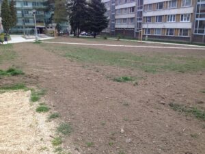 Nejednotná, většinou holá hliněná plocha s řídkou trávou je obklopena středně vysokými bytovými komplexy a několika stromy. Podél levé strany snímku vede štěrková cesta.