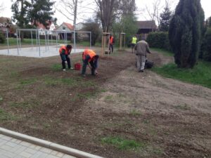 Několik pracovníků v oranžových bezpečnostních vestách sází nebo upravuje půdu v parku poblíž mladých stromků, zatímco další osoba používá nářadí na trávu. V pozadí jsou vidět bytové komplexy a vybavení dětského hřiště.