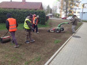 Čtyři pracovníci v ochranných vestách a teplém oblečení připravují půdu u chodníku poblíž bytové komplexy; jeden z nich obsluhuje rotační kypřič, zatímco druhý používá válec. V pozadí jsou vidět domy a bytové domy.