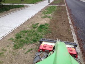 Pohled ze sedadla vozidla na údržbu zeleného trávníku, který ukazuje nejednotný travnatý pás s několika plevely mezi dlážděným chodníkem a silnicí v blízkosti bytové komplexy. Chodník vede souběžně se silnicí.