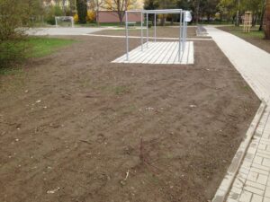 Plocha holé hlíny s řídkými rostlinami obklopuje kovovou konstrukci a dlážděné chodníky ve venkovním prostoru v blízkosti bytové komplexy, pravděpodobně v parku nebo na dětském hřišti. V pozadí jsou vidět stromy a tráva.