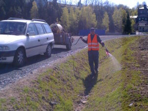 Osoba ve vestě s vysokou viditelností provádí chemické ošetření trávníku, stříká kapalinu z hadice do travnatého příkopu u silnice, poblíž je zaparkovaný bílý kombík s cisternou. V pozadí jsou vidět stromy a budovy.