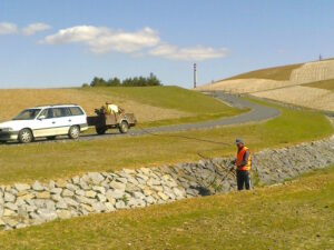 Osoba v bezpečnostní vestě stříká hadicí příkop obložený kameny a provádí chemické ošetření trávníku. Na nedaleké silnici v kopcovité travnaté krajině pod částečně zataženou oblohou parkuje bílé auto s přívěsem.