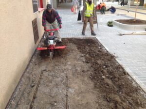 Dělník pracuje s červeným rotačním kypřičem na půdě vedle budovy, zatímco jiní pracují nedaleko na nově vydlážděném městském chodníku s terénními úpravami na pozadí moderních bytových komplexů.